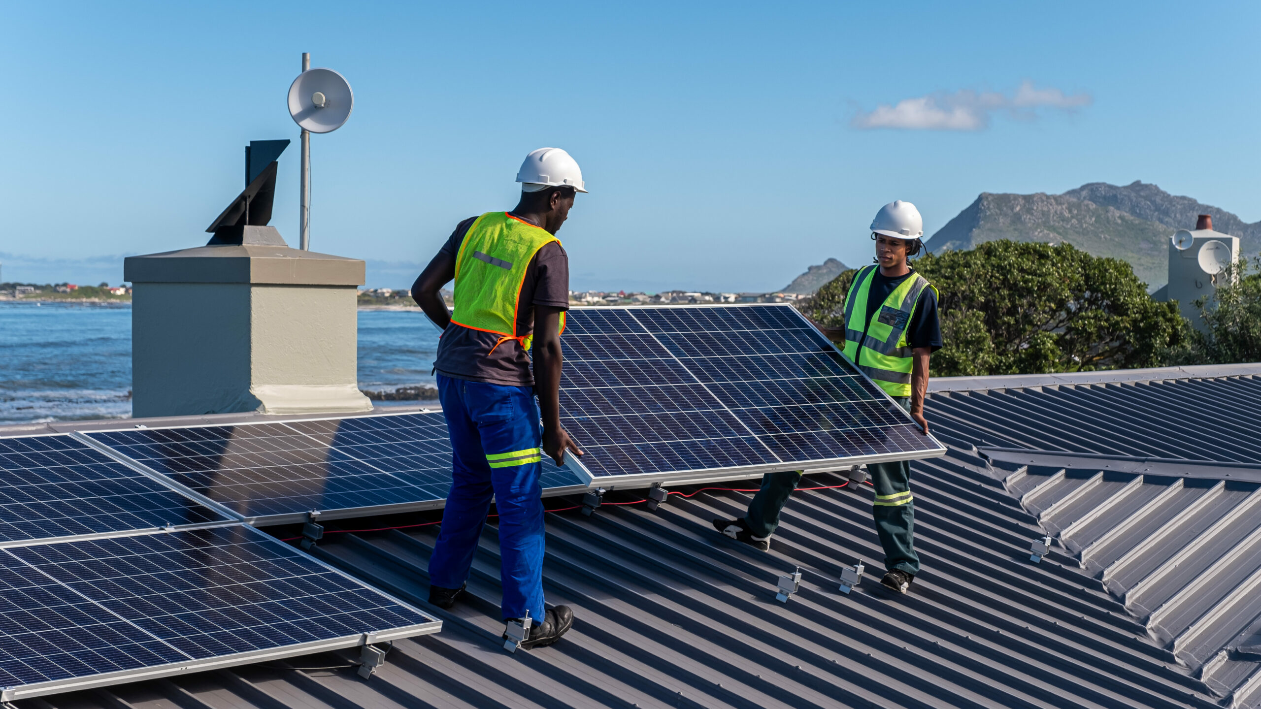 Two African men install solar panels neatly on a residential roof of a house near the ocean. Sustainable living.