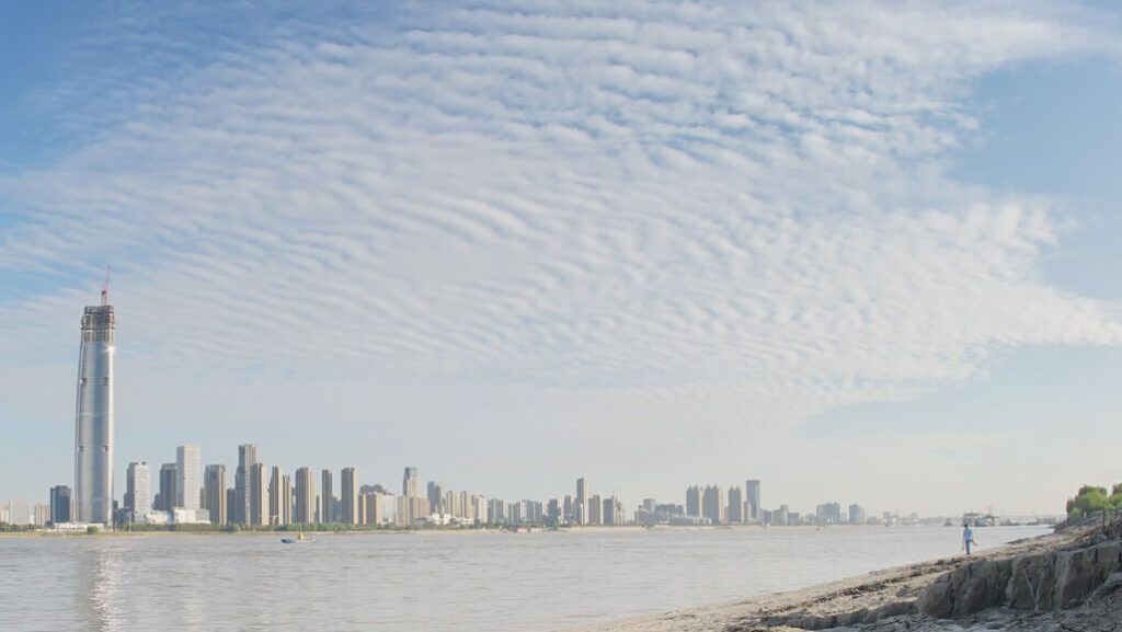 A person is stood on the bank of the Yangtze River in Wuhan, China, overlooking the city skyline.