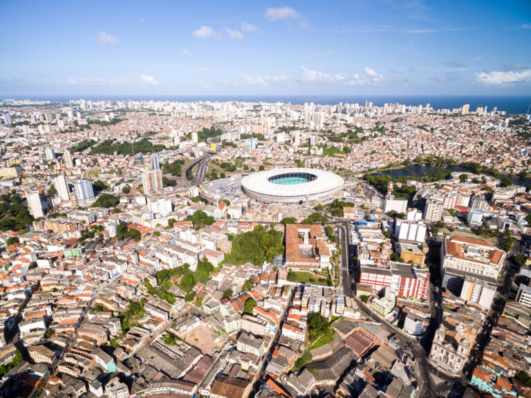 Photo of the city of Salvador, with many residential and commercial buildings, a park, and a stadium, with the ocean in the distance.