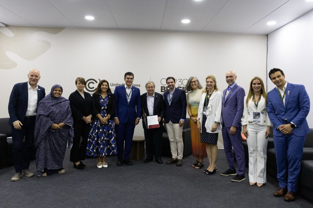 A group of 11 men and women in business and smart casual attire pose for a photograph in an indoor conference setting. A man in the center, wearing a gray suit, holds a white document. A wall behind them features logos for the United Nations and COP30 Brasil.